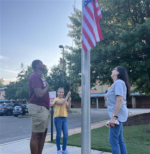 student raising flag in front of school as two adults salute